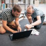 to mænd på knæ på gulvet, studerer intenst en skærm på en bærbarcomputer