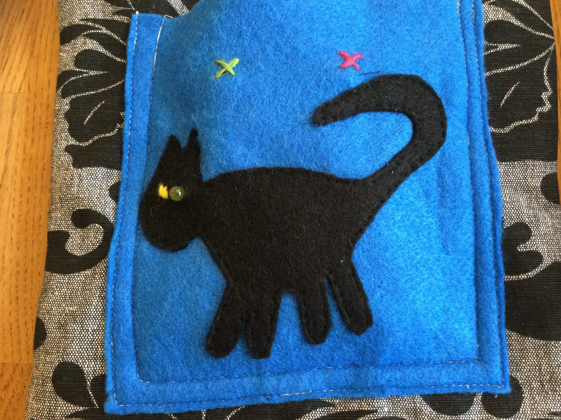 Taske med blå lomme med sort kat, der har dioder som øjne. Et grønt og et rødt kryds er syet og markerer, hvor der kan tændes og slukkes for dioderne